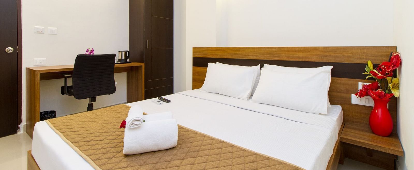 A hotel room including a double bed with brown bedding, a dark wardrobe, and a work desk and chair at Sanctum Suites Whitefield, Bangalore.