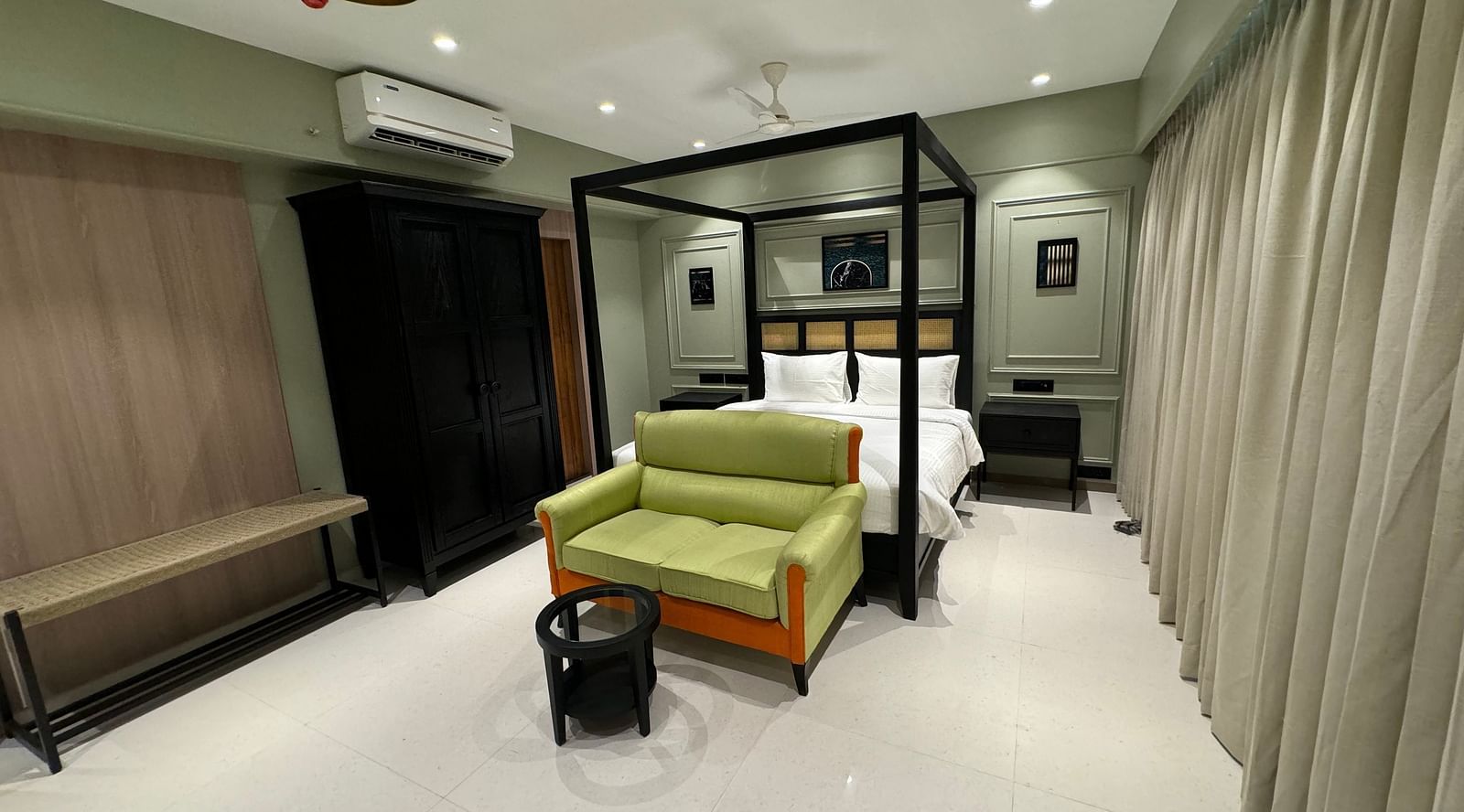 A hotel room with a black four-poster bed and white bedding, a green sofa, a black coffee table, a woven bench, and a black wardrobe, illuminated by ceiling lights and lamps.