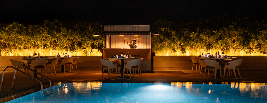 A tranquil nighttime view of the hotel's outdoor swimming pool, lit up with glowing lights, with a seating area surrounded by plants at The Residency Towers Rameswaram.