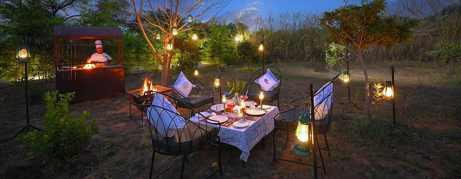 Private outdoor dining setup in a natural garden with cushioned chairs around a table adorned with lanterns and a live barbecue station, set against a serene twilight sky - Singhvilas, Ranthambore