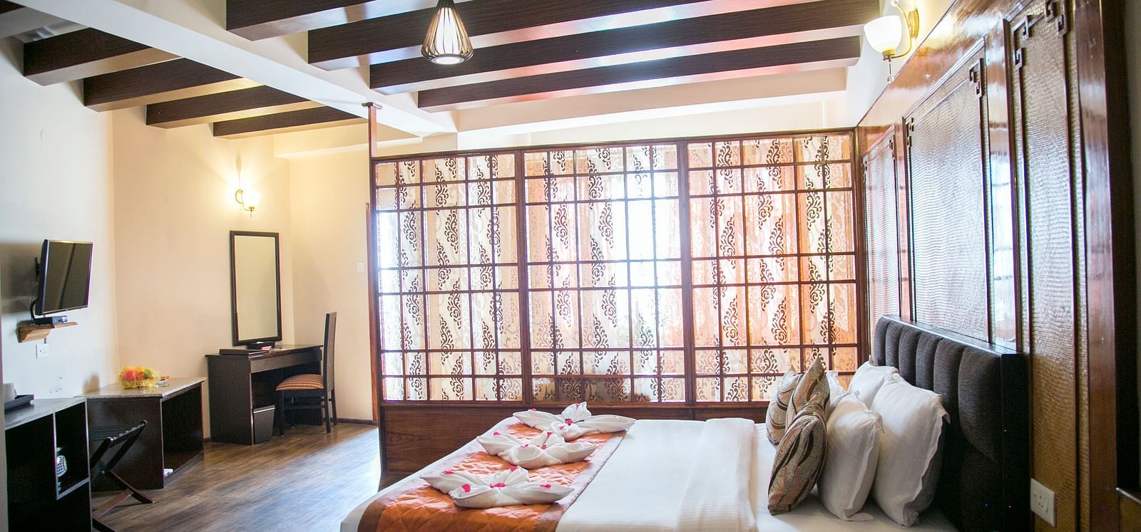 A room at New Orchid Lords Inn Gangtok contains a bed with pillows and towel swans, a bedside table with water bottles, a desk with a mirror and chair, and a TV.