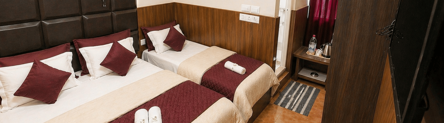 A double bed next to a single bed, with a wall-mounted TV and a wardrobe in the Deluxe Triple Room at BN Tourist Home, Ooty.