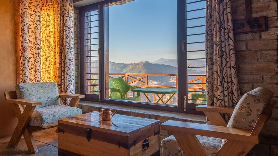 A tranquil interior view showcasing floral curtains, wooden furniture, and a stunning outdoor mountain landscape. - Blusalzz Homes The Himalayan Bungalow, Almora