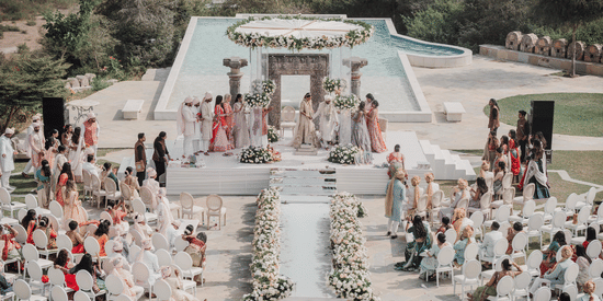 A wedding ceremony at the outdoor featuring as aisle decorated with flowers on either side, guests seated on either side and a centre stage where the rituals are taking place  with bride, broom, and their close family members.