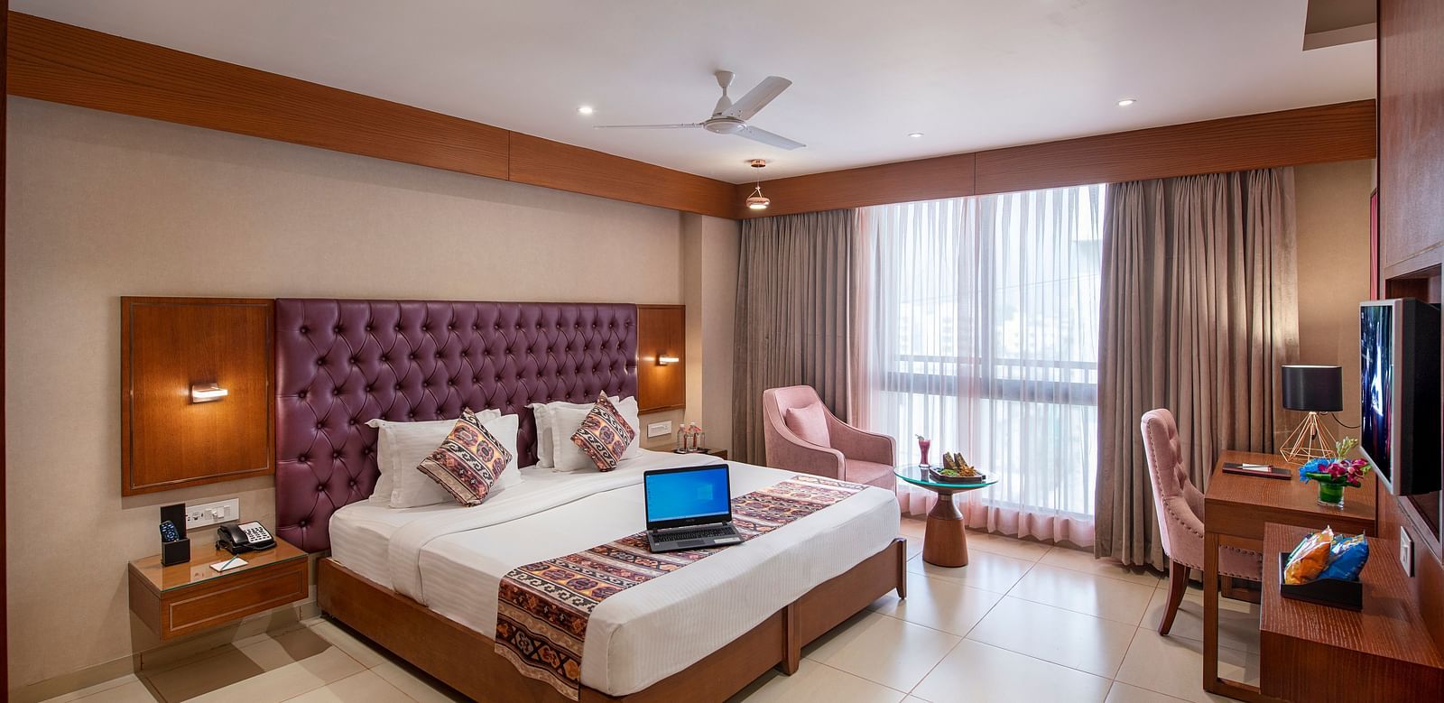 A well-lit hotel room with a large bed with a purple headboard, a work desk, and a seating area by the window - SK Lords Inn Ahmedabad