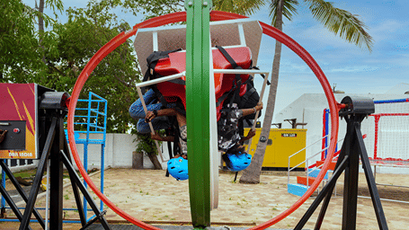 Two riders in blue helmets strapped into the red seats of the Gyro ride, inside a red and green ring frame on a steel platform at MGM Dizzee World, Chennai.