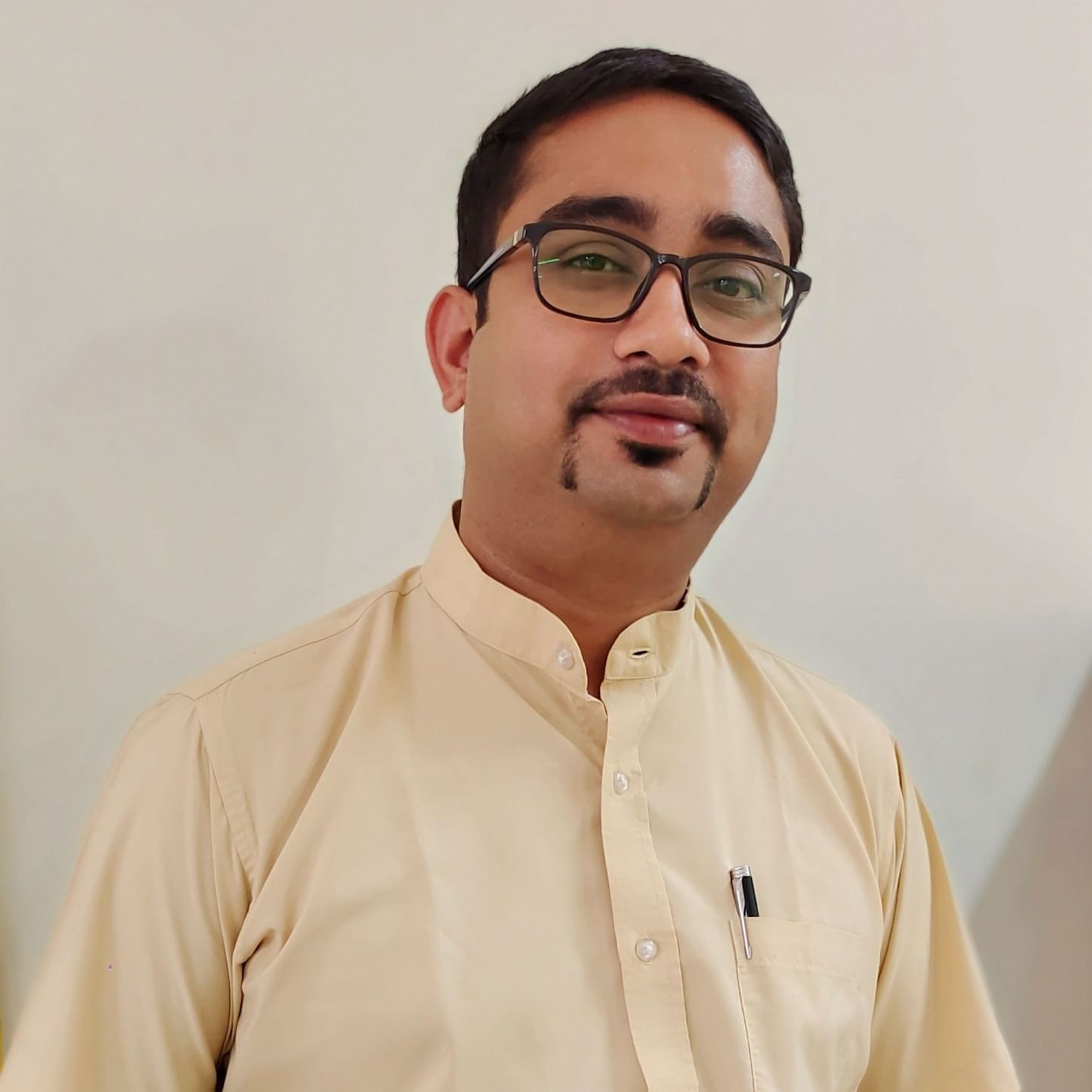 A man with glasses and a mustache, wearing a light-coloured collared shirt, smiles at the camera