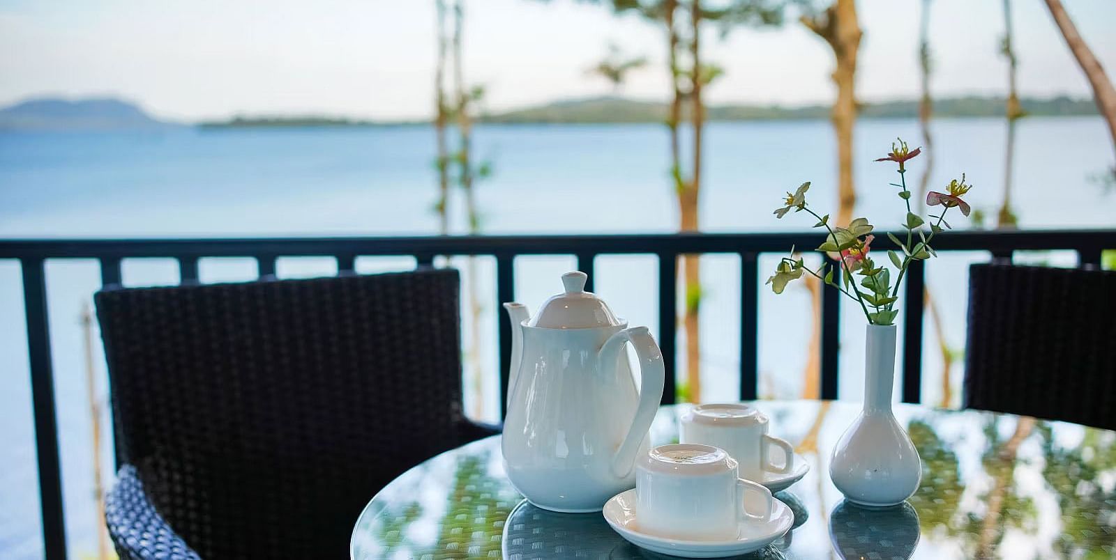 Ceramic water jugs and cups with saucers placed on a glass dining table with the backwaters at Coorg Jungle Camp Backwater Resort, Kushalnagar in the backdrop.