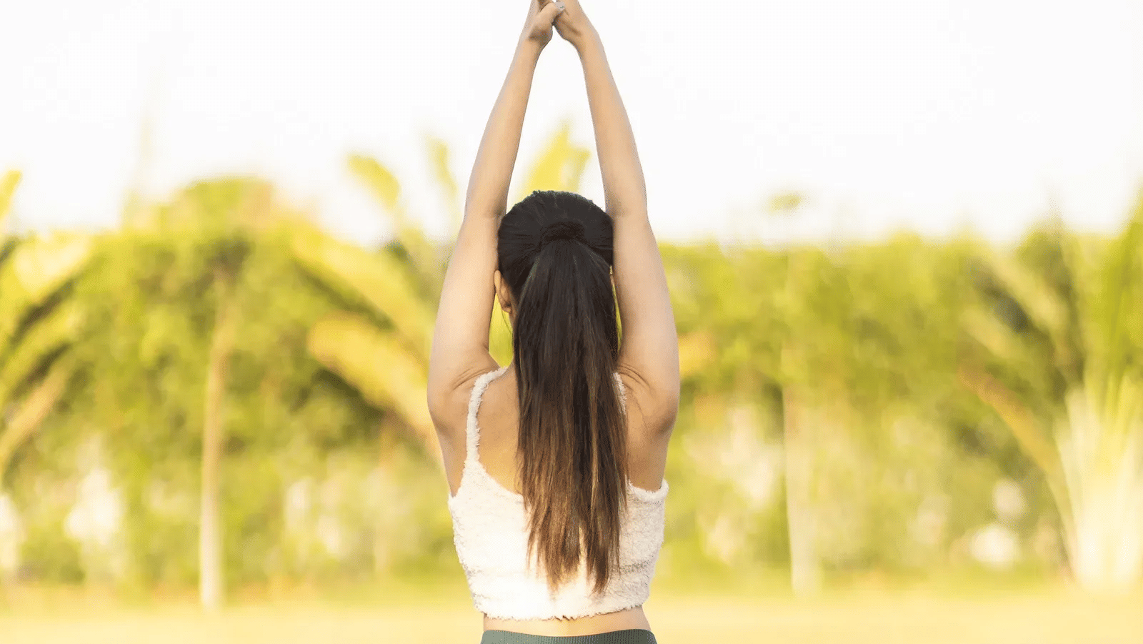 Back view of a woman wearing sports gear and in a yoga posture with blurry background of grass lawn and trees -Daksh Eden Greenz, Sasan Gir