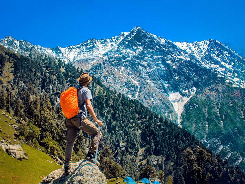 A person on treks near Dehradun with an orange backpack stands on a rocky mountain ridge, overlooking snow-capped Himalayan peaks beneath a clear blue sky.