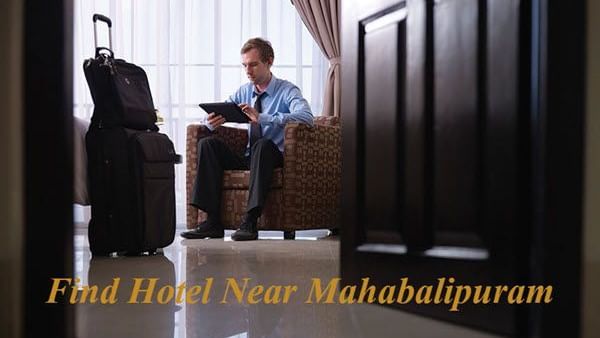 A man sitting on a sofa chair, with a suitcase and a small bag, beside a large window with the text “Find Hotel Near Mahabalipuram” showing.