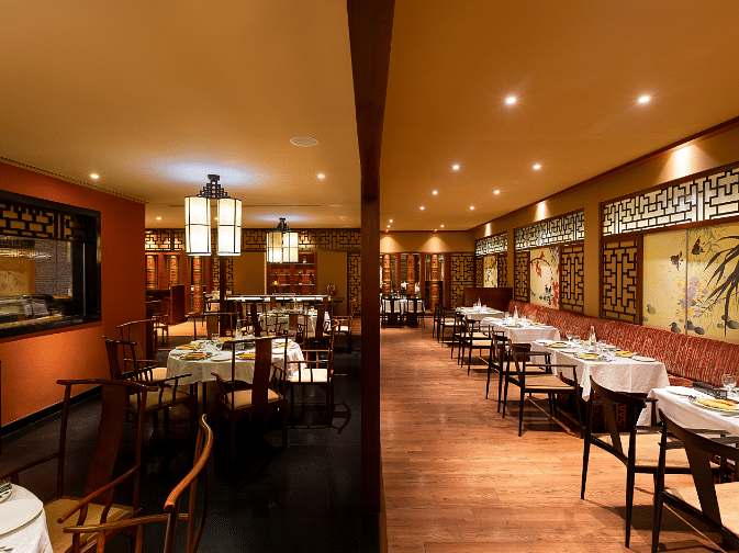 A well-lit restaurant with a long dining table, chairs, and a bar counter in the background.