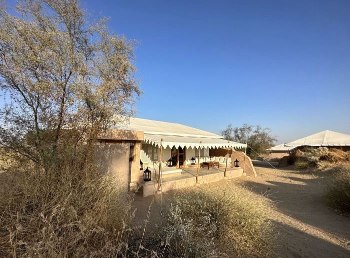 A luxury desert tent with an extended canopy stands amid a dry landscape with sparse trees, under a clear sky - MANVAR Resort & Desert Camp