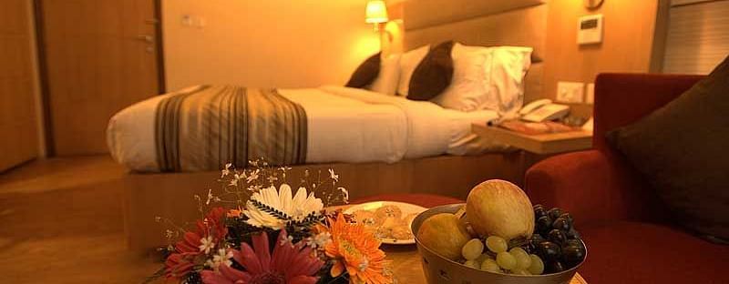 A fruit bucket filled with assorted fresh fruits and a colourful flower vase placed  beside a bed  at The Manor, Bareilly