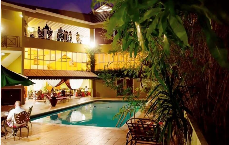 An outdoor view of a hotel at night, featuring a swimming pool surrounded by palm trees and illuminated buildings | Altamont Court, Jamaica