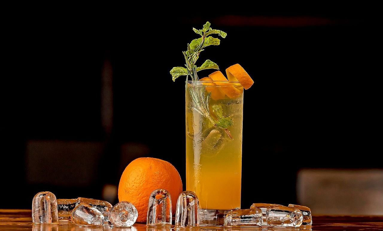 A refreshing cocktail garnished with mint leaves and orange slices, served in a tall glass with ice cubes and a whole orange on a wooden surface against a dark background.
