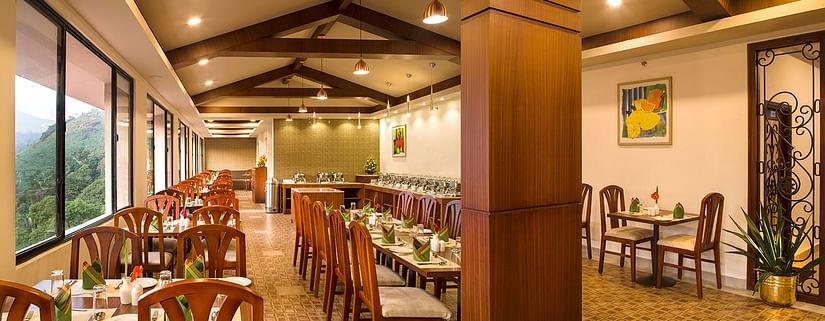 Restaurant interior showing a long row of tables and chairs. The room has large windows, wooden pillars, and exposed wooden beams - Abad Copper Castle, Munnar
