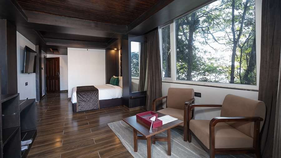 A sweeping shot of the room, showcasing the bed, cosy seating area with two chairs, and a table beside a grand window.