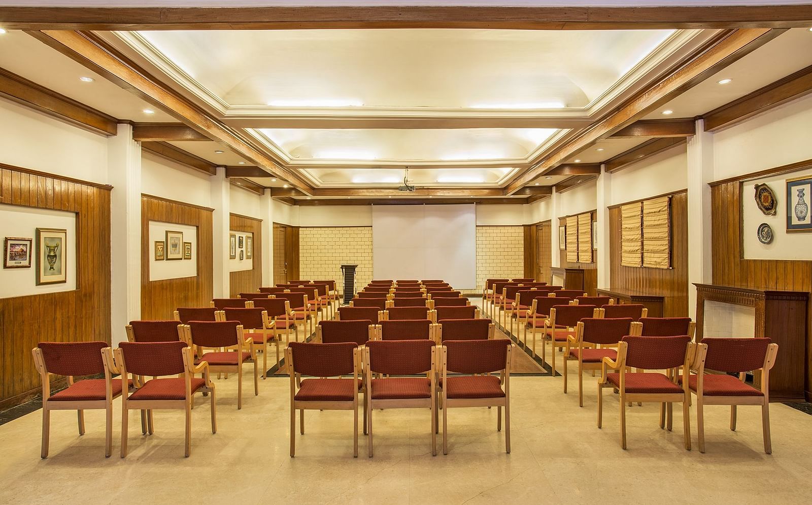 A spacious conference room with rows of chairs facing a projection screen. The room has high ceilings, wooden paneling, and framed artwork on the walls. The overall ambiance is professional and formal - the naini retreat, nainital.