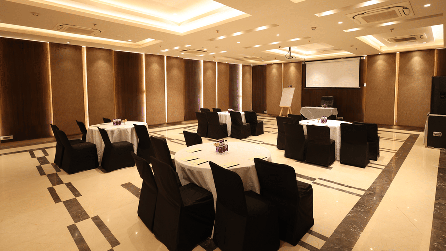 Banquet hall at Hotel Saket 27, New Delhi with round tables, black chair covers, projector screen and warm ceiling lighting.