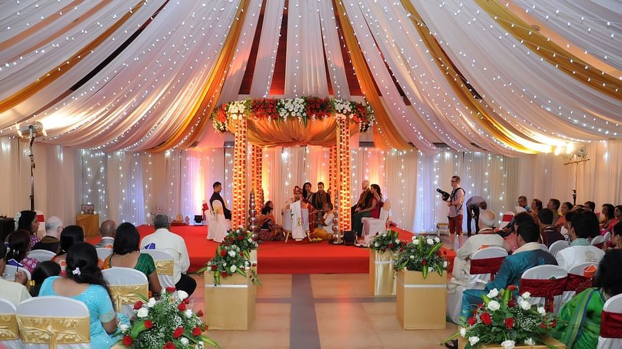 An image of an indoor wedding hall with a floral decorated mandap at the center, guests seated around, and lights hanging from the ceiling, creating a festive atmosphere