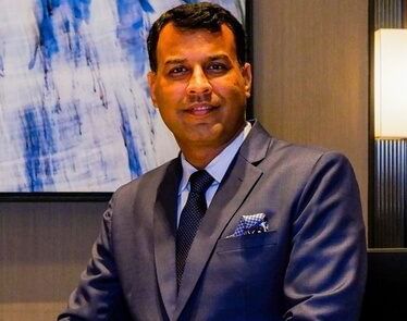 Professional headshot of a man in a dark suit and tie, seated in an indoor setting with abstract blue artwork on the wall.