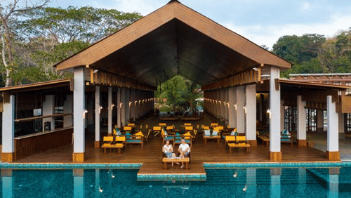 Poolside pavilion at Symphony Oceanus Bay Beach Resort & Spa with a peaked wooden roof, white pillars, teal loungers, and guests beside the pool at dusk.