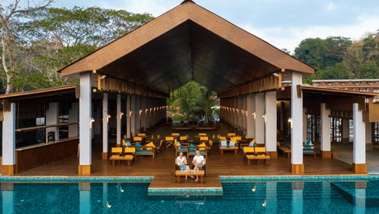 Poolside pavilion at Symphony Oceanus Bay Beach Resort & Spa with a peaked wooden roof, white pillars, teal loungers, and guests beside the pool at dusk.