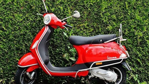 A vibrant red Vespa scooter is parked against a lush green hedge.