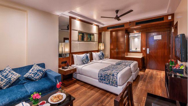 A hotel room is furnished with two single beds, a sofa, a table with snacks, a television, and wooden flooring. 