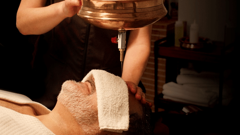 A close-up shows a man lying down with a towel on his forehead, receiving a shirodhara treatment with warm oil pouring from a brass pot at YO1 Longevity & Health Resorts