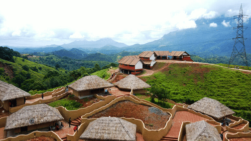 Aerial view of tribal huts with thatched and tiled roofs built on hillsides against a backdrop of green mountains and a cloudy sky.