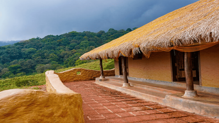 Close-up of a traditional hut with a thatched roof, supported by wooden posts, featuring a tiled porch and a curved mud wall, overlooking a forested hill..