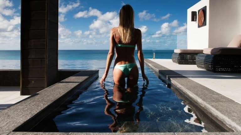 A woman in a swimsuit stands in a narrow pool, looking out at a stunning turquoise ocean under a bright blue sky with clouds.