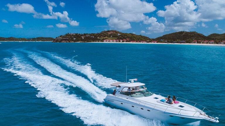 White luxury yacht cruising through vibrant blue waters, leaving a foamy wake behind, with a tropical coastline and lush green hills under a partly cloudy sky in the background.