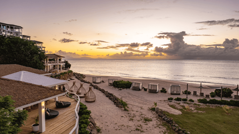 Sunset over a sandy beach with lounge areas and oceanfront villas, palm trees silhouetted.