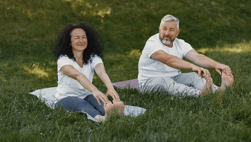 An older couple sits on yoga mats in the grass, smiling and stretching outdoors, enjoying a peaceful wellness moment in nature