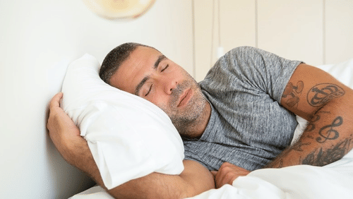 A man lies peacefully on a white pillow in a bed with his eyes closed
