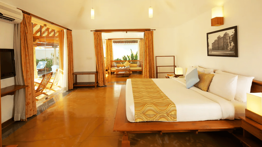 A well-lit hotel room featuring a large bed with a gold-patterned runner and a view into an adjacent seating area | Le Pondy