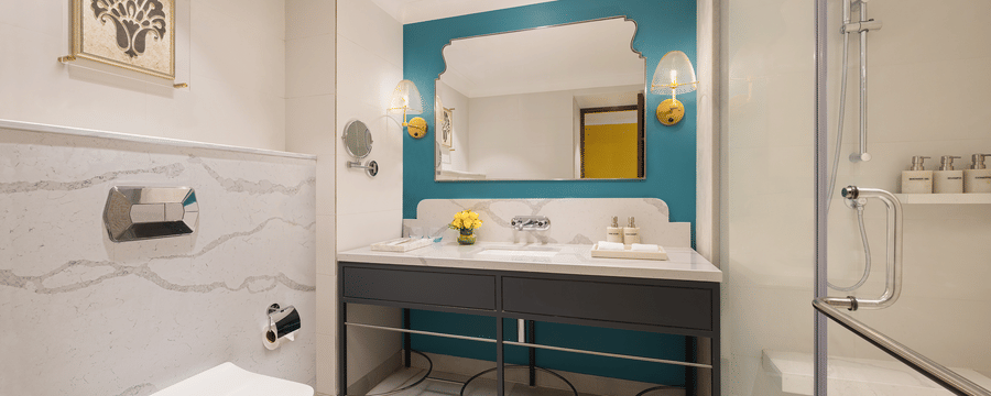 A stylish bathroom with a white toilet, a modern sink with a marble countertop and a large mirror framed by a teal wall, sconce lighting, a glass-enclosed shower, and a framed artwork on the wall - Aloha On the Ganges.