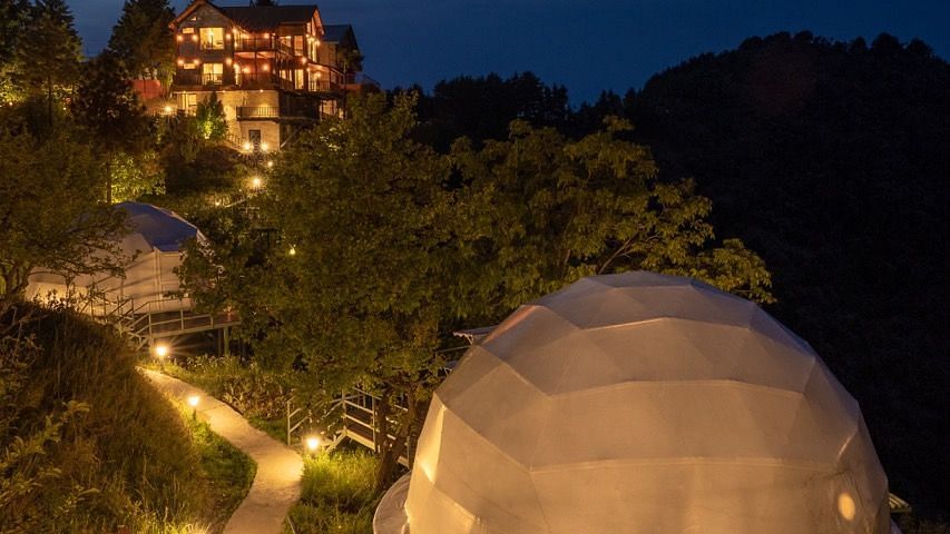 An image of a well lit dome and a facade at a distance and the pathways are lit with lights too -  Avalon Cottages, Kanatal