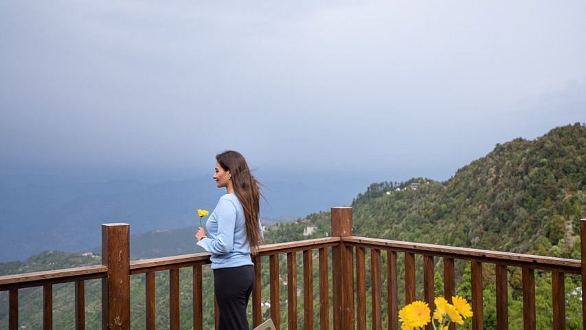 An image of woman enjoying the view from avalon cottages along with a chair and table being around | Avalon Cottages, Kanatal
