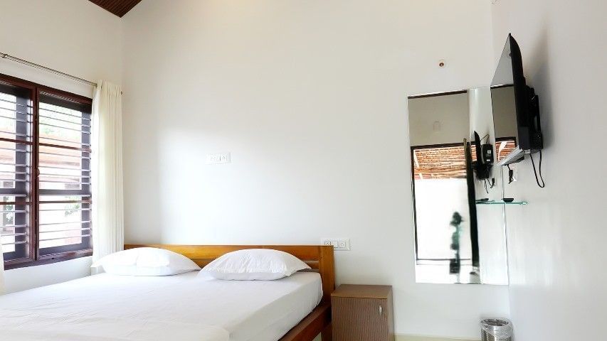 A spacious deluxe room at Bhuthanakadu Retreat, featuring a queen-sized bed with crisp white linens, a wooden headboard, and a high, pitched wooden ceiling with a fan.