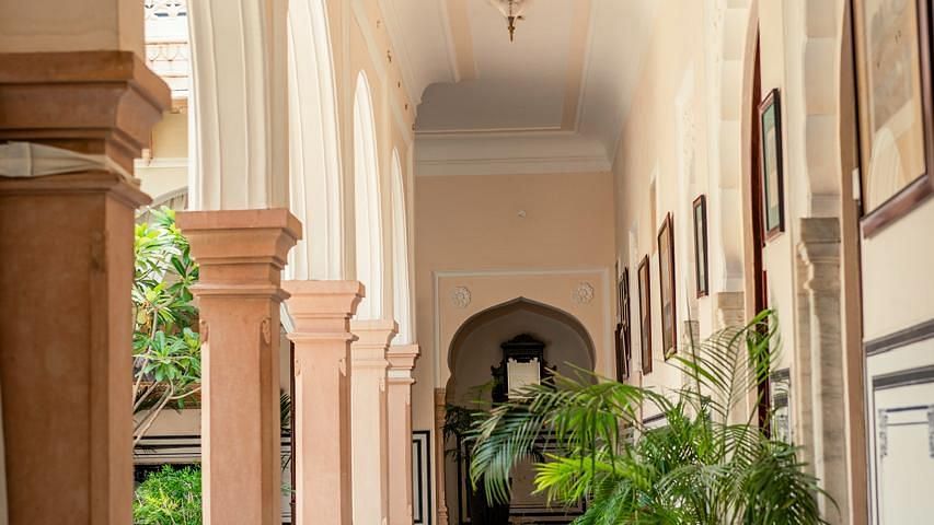 A sunlit corridor with monochrome flooring, arched walls, and potted plants along the walkway - Khas Bagh, Jaipur.