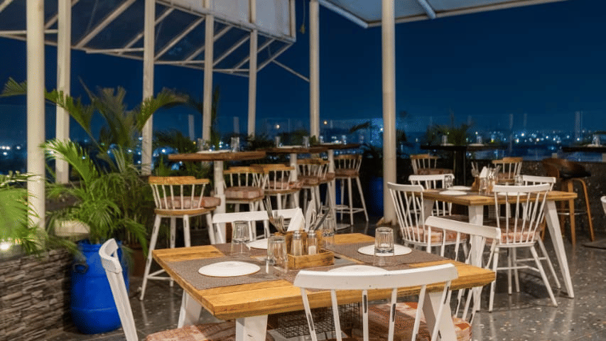 Outdoor seating area of Papaya Tree Hotels with neatly arranged wooden tables and chairs overlooking the open view under a covered roof at the Nazaara Restaurant.