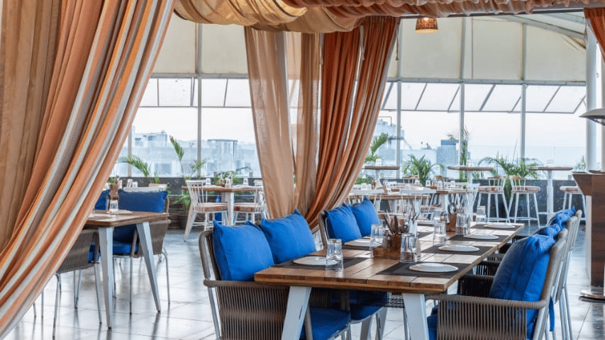 Indoor dining setup of Nazaara Restaurant at Papaya Tree Hotels , featuring draped curtains, cushioned seating, and wide windows providing a scenic backdrop.