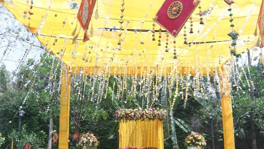A decorated outdoor wedding setup at Tisya Farms, Gurgaon with yellow drapes, flags, and a ceremonial stage on a green carpet.