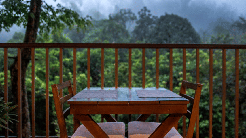 A balcony with a simple wooden table and chairs, overlooking misty hills.- Abad Brookside Lakkidi, Wayanad