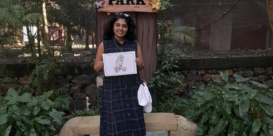 A woman standing in front of the EsselWorld Bird Park signboard, holding a sketch, surrounded by plants and trees.