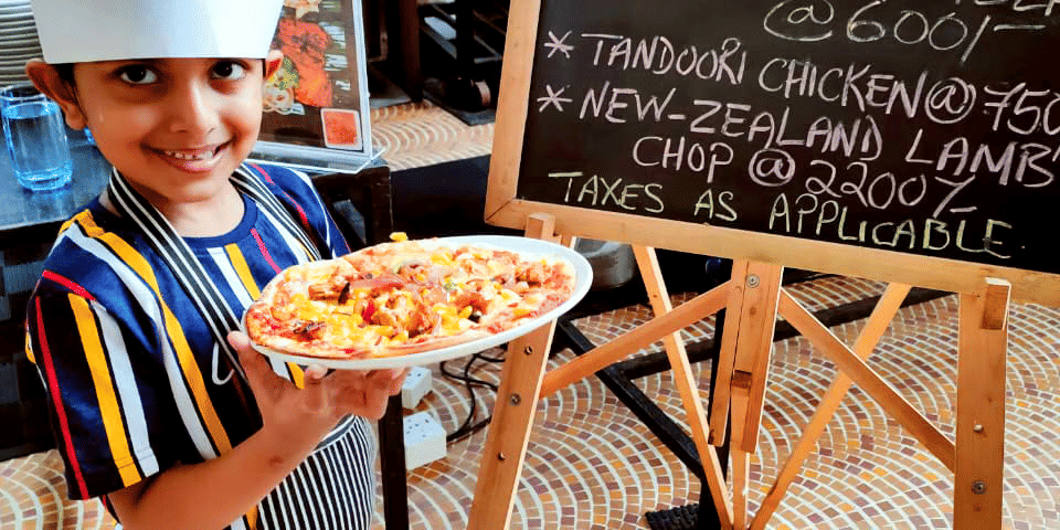 A kid wearing a chef hat, holding a plate of pizza with the chef in the background at Caravela Beach Resort